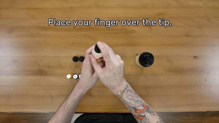 An image of the artist placing their index finger over the tip and cap of a sealed batter pen with batter and unmixed coloring inside of it that is in their grasp. The image reads, "Place your finger over the tip,"