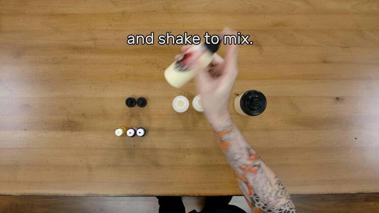 An image of the artist, with the batter pen firmly grasped with their index finger over the tip of the bottle, beginning to vigorously shake the batter pen to mix the red gel coloring inside of it into the batter itself. The image reads "and shake to mix."