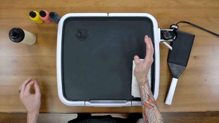 An image of the dancakes pancake art griddle, and the artist is resting their drawing hand on a folded-over white washcloth, demonstrating how artists can use folded hand towels to lend their drawing hand more stability without risking a burn.