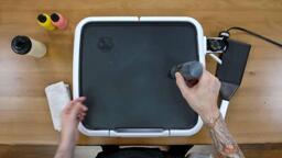An image of the dancakes pancake art griddle, over which the artist is gently gripping a black batter pen, demonstrating how suction can be used to prevent batter from pouring out automatically - a technique called 'squeeze control'.