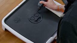 An image showing the black outlines of a heart eyes emoji being drawn onto the dancakes griddle with pancake batter.