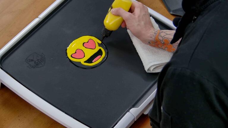 An image showing a heart eyes emoji pancake design having its yellow face colored in, the red eyes and mouth already having been colored in.