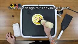 An image showing the artist drizzling a zig-zag of plain batter across the back of the heart-eyes emoji design. The image reads "'Back' your design to fill in any missed spots."