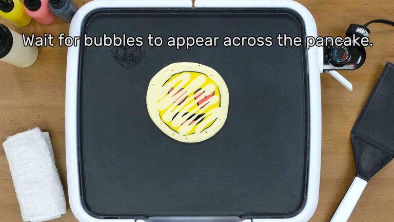 An image of a heart-eyes emoji pancake design that has been bordered and backed, beginning to bubble on the griddle as it cooks. The image reads "Wait for bubbles to appear across the pancake."