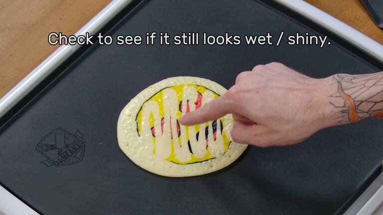 This image shows the artist pointing to the back of the cooking pancake art design to point out how the texture of the pancake is slowly changing from shiny to matte. As it cooks, it becomes less 'wet'. The image reads "Check to see if it still looks wet / shiny."