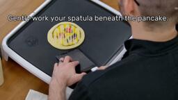 This image shows the artist, holding the spatula, starting to slide the spatula under and around the pancake design, which is gently stuck in place. The image reads "Gently work your spatula beneath the pancake."