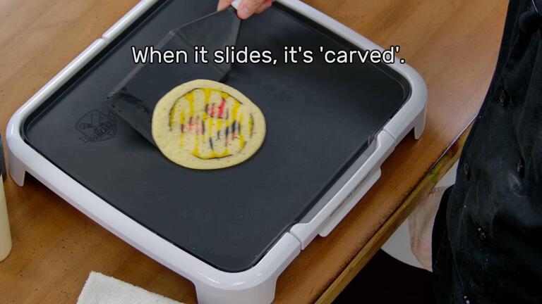 This image shows the artist, holding the spatula, sliding the pancake design around (the design appears with a motion blur in the photo). The image reads "When it slides, it's 'carved'."