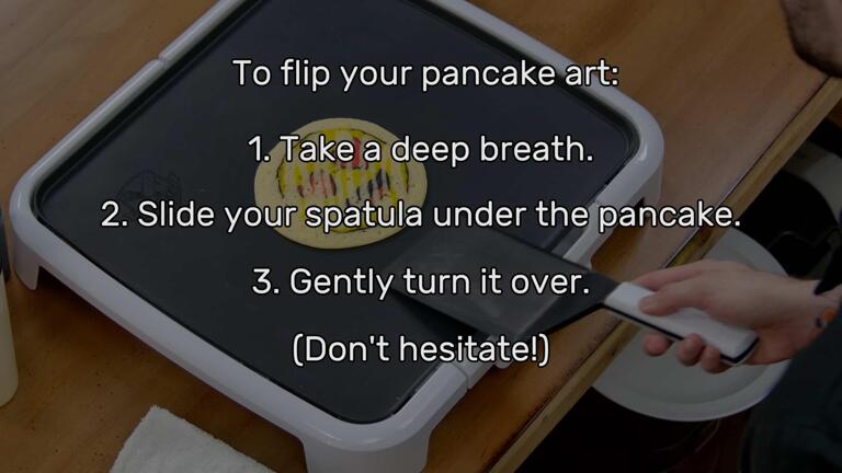 An image of a spatula about to slide under the heart eyes emoji pancake art design. The image has been faded out to reveal a list of text that reads: "To flip your pancake art: 1. Take a deep breath. 2. Slide your spatula under the pancake. 3. Gently turn it over. (Don't hesitate!)"
