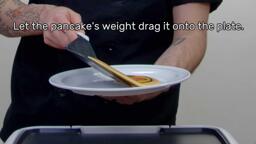 An image of the heart eyes emoji pancake design being plated, with the text "Let the pancake's weight drag it onto the plate."
