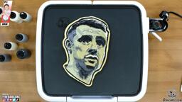 An image of a pancake art portrait of social media personality Gary Vaynerchuck, rendered in grayscale and pictured resting on the dancakes griddle from a top-down viewing angle. The warm brown wood of the countertop and the multicolored batter pens adorn the edges of this simple image.
